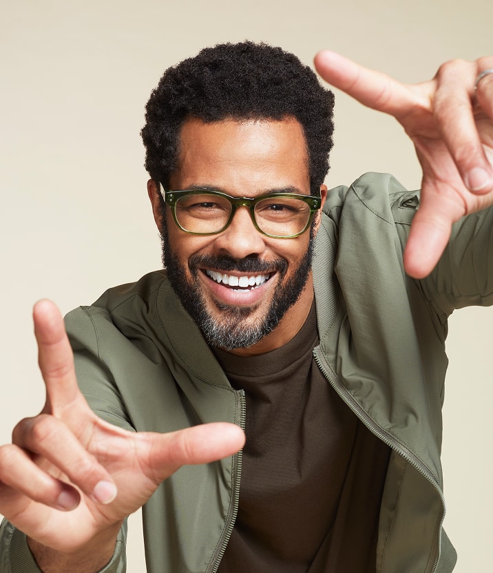 A smiling man with facial hair wearing a green casual jacket and Zenni Rectangle Glasses, framing his face with his hands against a beige background.