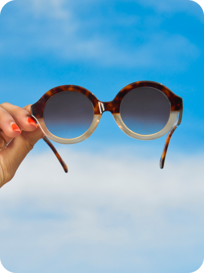 A woman’s hand holds up a pair of Zenni round tortoiseshell sunglasses against a blue sky background.