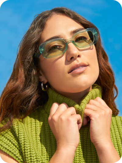 A young woman with long brown hair wearing translucent green Zenni rectangle glasses and a green turtleneck against a bright blue background.