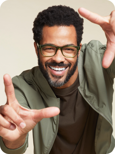 A smiling man with facial hair wearing a green casual jacket and Zenni Rectangle Glasses, framing his face with his hands against a beige background.