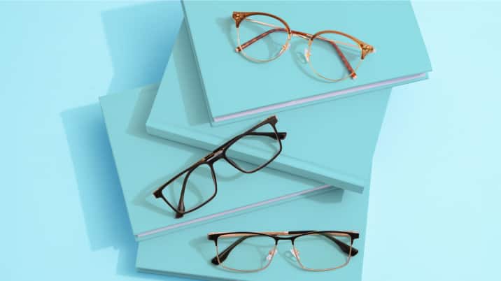 Three pairs of eyeglasses displayed on a stack of light blue books. The glasses include a round gold frame, a rectangular black frame, and a semi-rimless black frame.