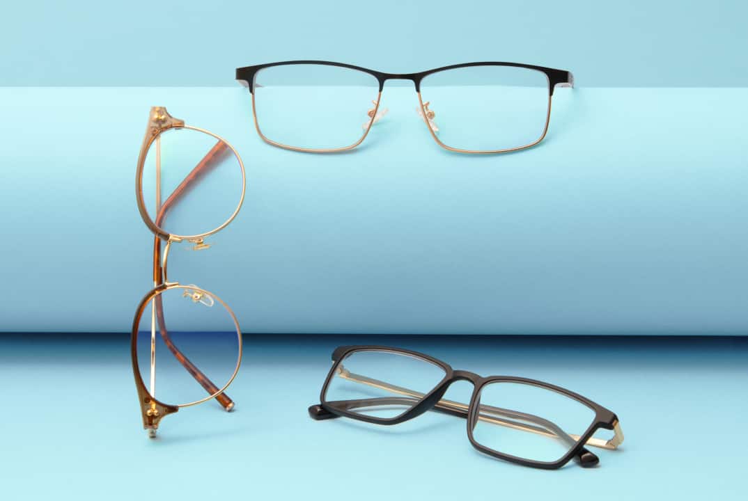 Three pairs of eyeglasses arranged on a blue background. The pairs include a round gold frame, a rectangular black frame, and a semi-rimless black frame.