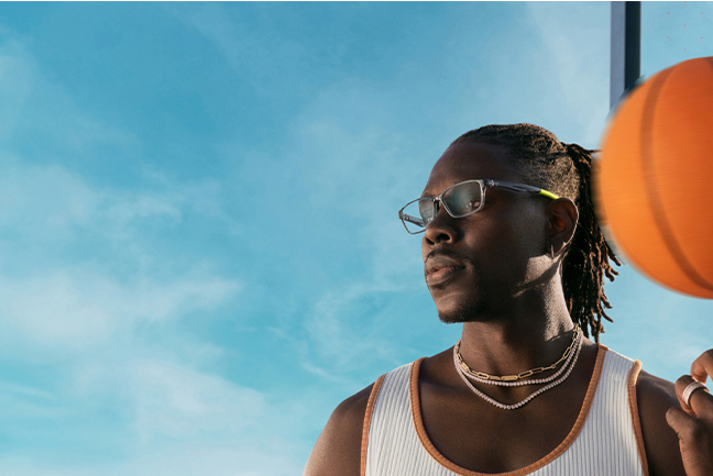 Jrue Holiday wearing layered gold and silver necklaces, clear rectangle glasses with a gold detail on the arms. He holds a basketball in motion while standing against a bright blue sky.