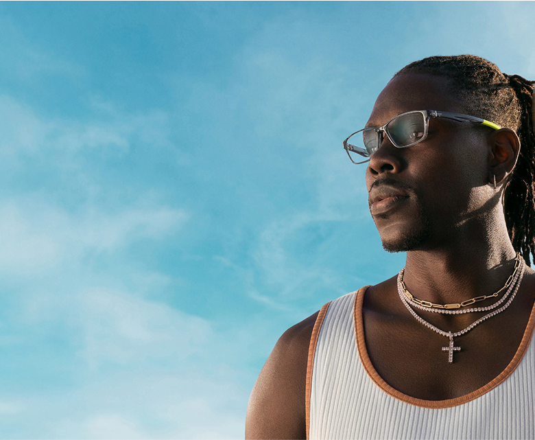 Jrue Holiday wearing layered gold and silver necklaces, clear rectangle glasses with a gold detail on the arms. He holds a basketball in motion while standing against a bright blue sky.