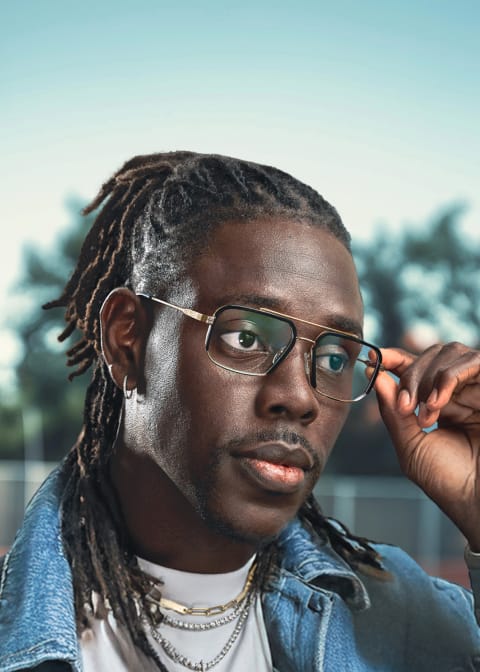 Boston Celtics point guard Jrue Holiday wears “Paragon” aviator optical glasses from his collection with Zenni while looking away from the camera wearing a denim jacket accessorized with silver earrings and silver chain necklace.