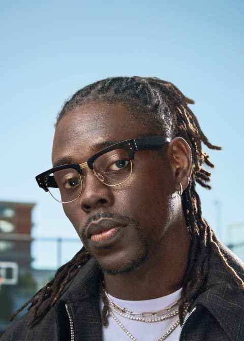 Boston Celtics Point Guard Jrue Holiday wears black eyebrow “Vanguard” optical frames from his Zenni collection, looking into the camera in a black jacket and silver chain, with a blue sky behind him.