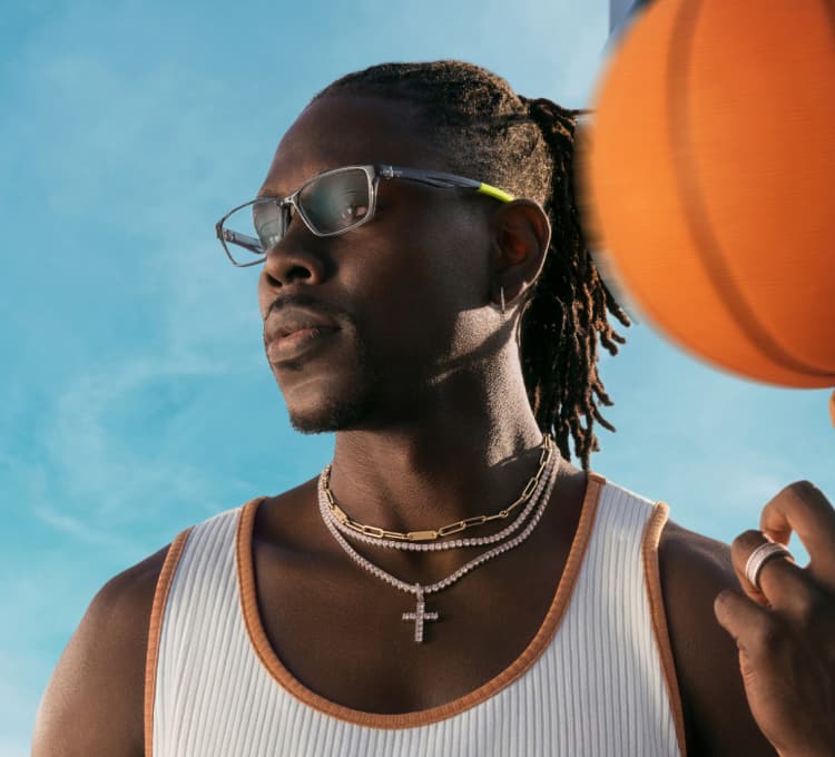 Boston Celtics Point Guard Jrue Holiday wears gray rectangle “DefenderX” frames from his Zenni eyewear collection. He stands against a blue sky, spinning a basketball on one finger while gazing outward.