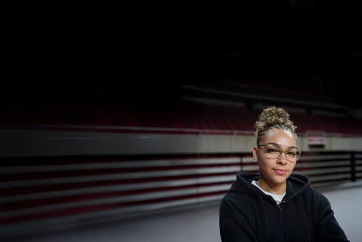 A portrait of a woman with her hair in a high bun wearing clear, light-green cat-eye eyeglasses, posed in a stadium with rows of red seating blurred in the background.