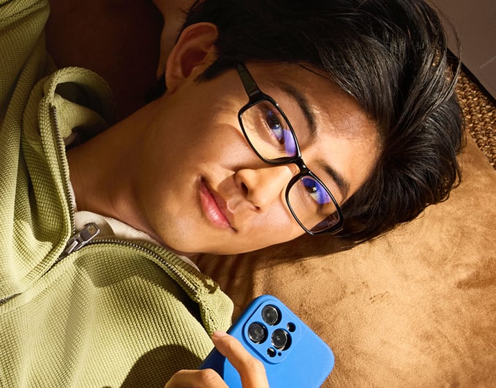 Man relaxing on a pillow while holding a smartphone and wearing black rectangular blue-light blocking eyeglasses.