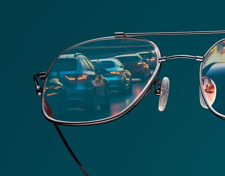 Metallic aviator eyeglasses with lenses reflecting nighttime highway traffic lights against a teal background.
