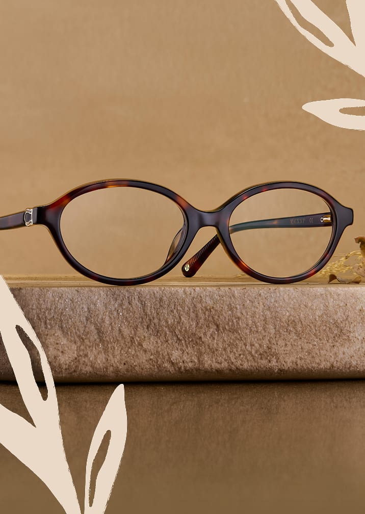 Tortoiseshell oval eyeglasses displayed on a stone pedestal against a warm beige background with leaf line art.