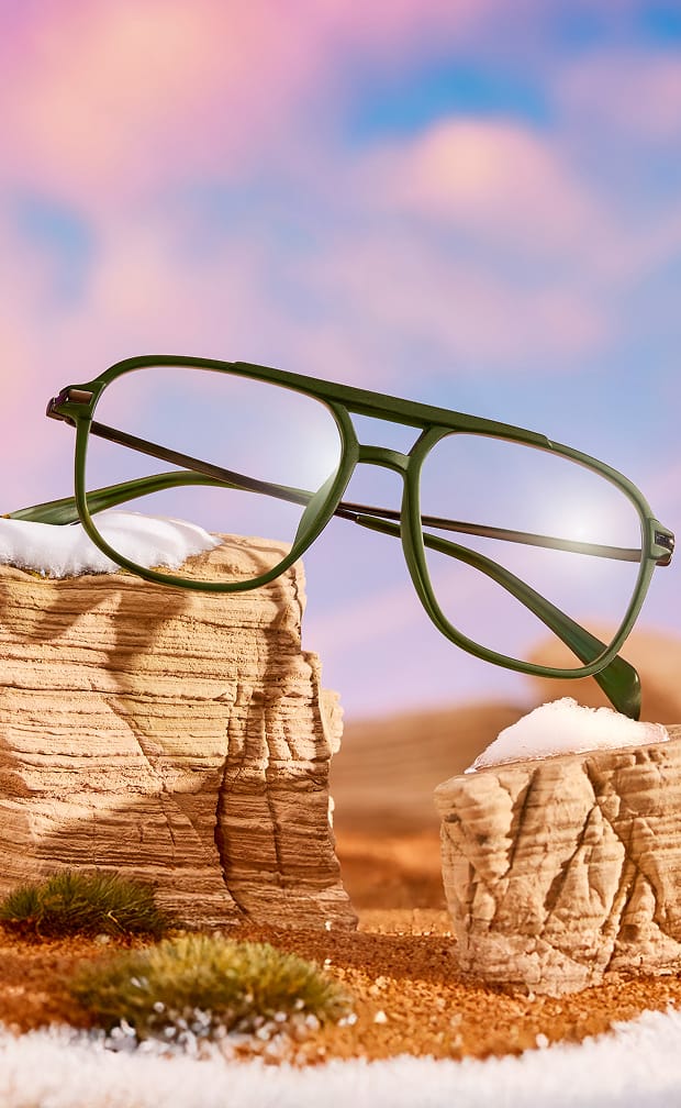 Olive green aviator eyeglasses balanced on desert rock formations against a soft pastel sunset sky.
