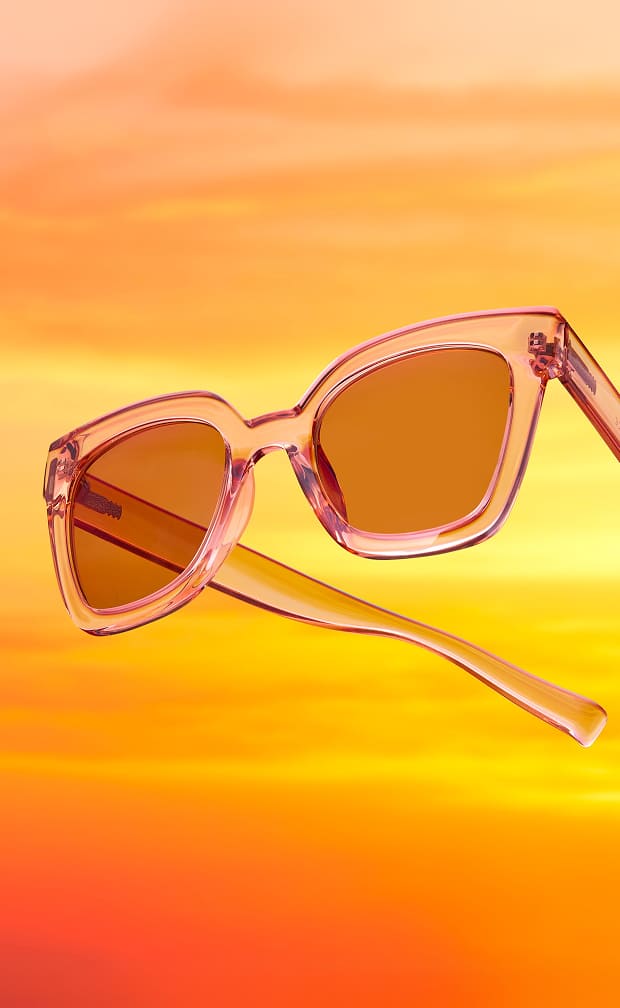 Translucent peach cat-eye sunglasses floating against a vibrant orange and yellow sunset background.