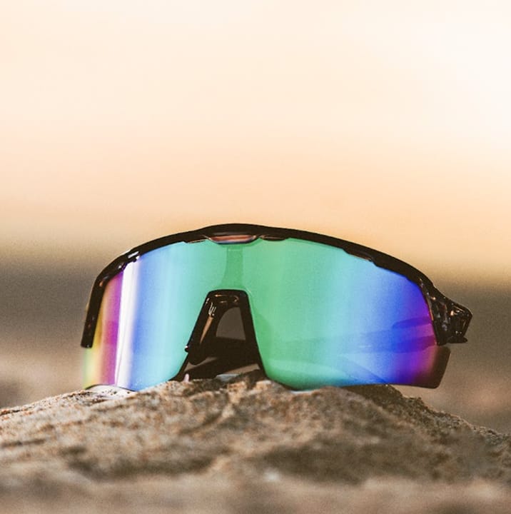 Text that says: ‘Maximum Ventilation.’ Kristen & Taryn love the cool views courtesy of premium airflow. Sunglasses with multicolored lenses on a sandy surface against a sunset background. 'Shop now' button below the quote.