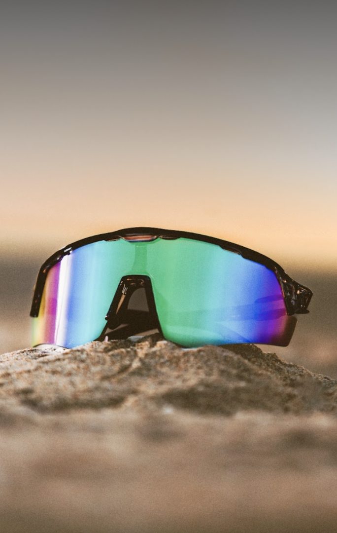 Text that says: ‘Maximum Ventilation.’ Kristen & Taryn love the cool views courtesy of premium airflow. Sunglasses with multicolored lenses on a sandy surface against a sunset background. 'Shop now' button below the quote.