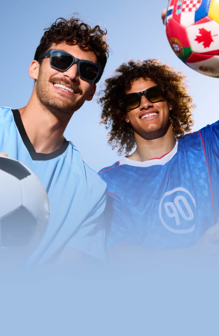 Dos hombres sonrientes usando camisetas deportivas y gafas de sol oscuras miran hacia la cámara, flanqueados por un balón de fútbol clásico y otro con estampado de banderas del mundo bajo un cielo azul despejado.