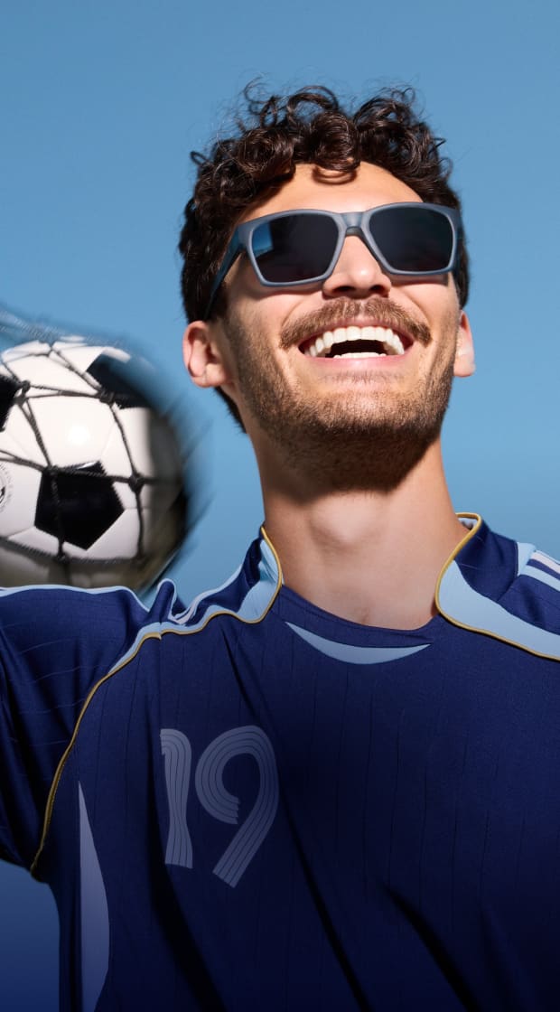 Primer plano de un hombre sonriente con cabello oscuro rizado y bigote usando lentes de sol cuadrados grises y una camiseta azul marino, mientras sostiene un balón de fútbol en una red sobre un fondo de cielo azul sólido.