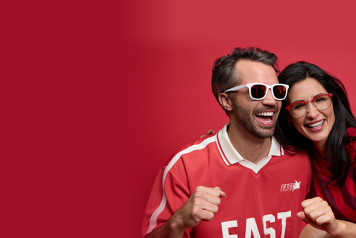 Two adults wearing Zenni eyewear and red soccer-inspired outfits, smiling against a red background.