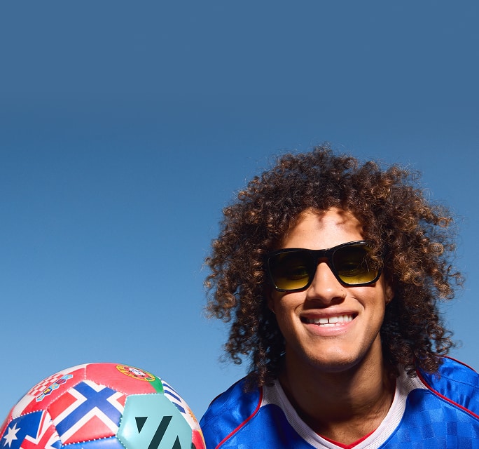Smiling person with curly hair wearing a blue soccer jersey and black sunglasses with yellow-tinted lenses, posing next to a soccer ball decorated with global flags against a solid blue sky.