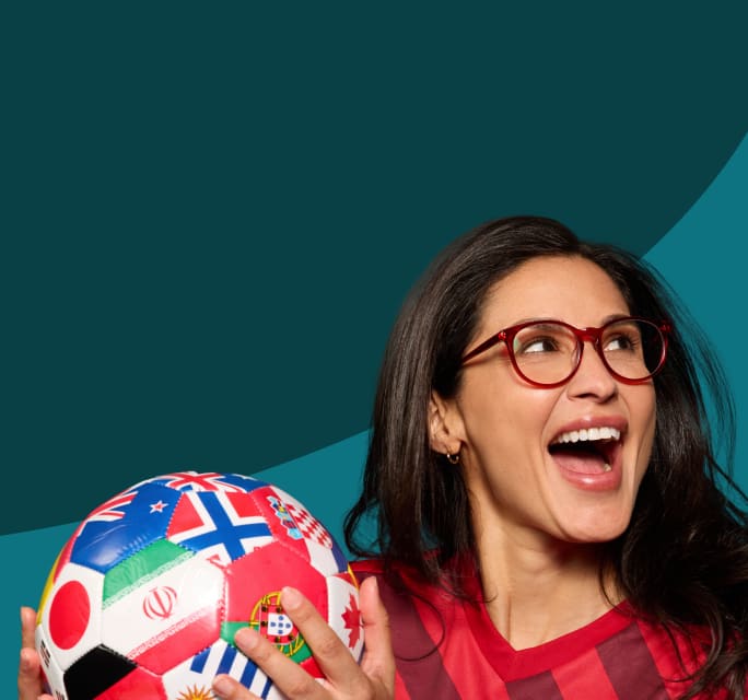 Enthusiastic woman looking up and laughing while wearing red round eyeglasses and a red striped soccer jersey, holding a soccer ball patterned with multi-national flags against a dark teal background.