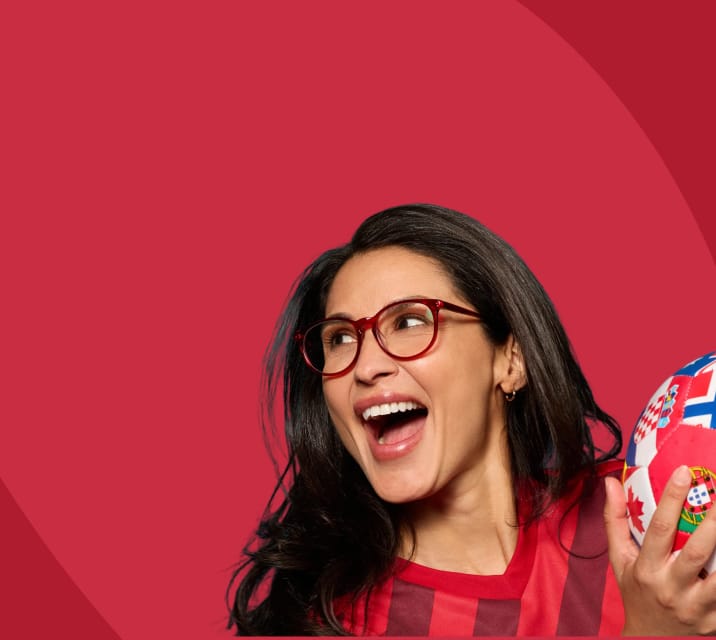 Excited woman looking to the side and laughing while wearing red round glasses and a striped red jersey, holding a multi-national flag patterned soccer ball against a vibrant red background.