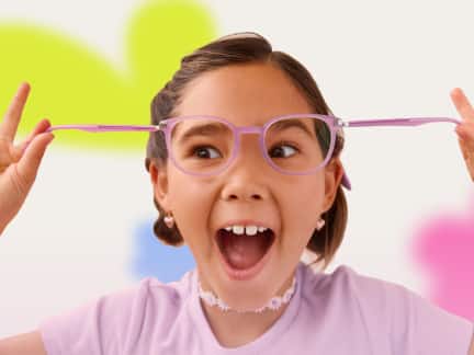 A young girl with pigtails excitedly stretches her pink glasses outward, wearing a lavender shirt and floral choker, with colorful blurred shapes in the background.