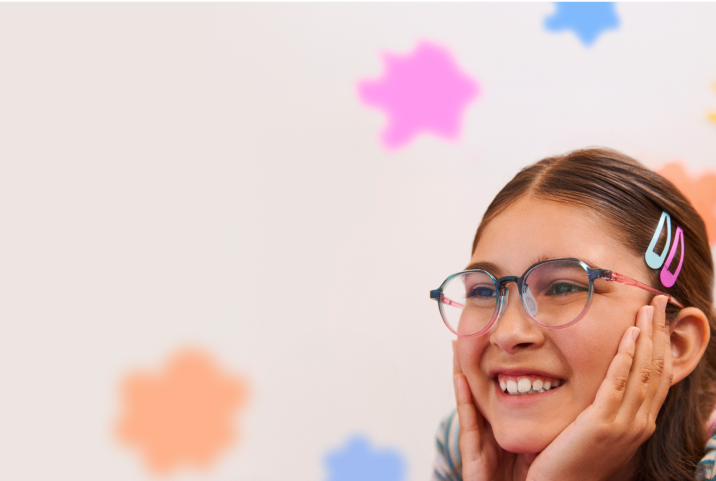 Girl with long brown hair and colorful hair clips wearing round pink-and-teal eyeglasses, resting her face in her hands and smiling, with soft orange and blue flower shapes blurred in the background.