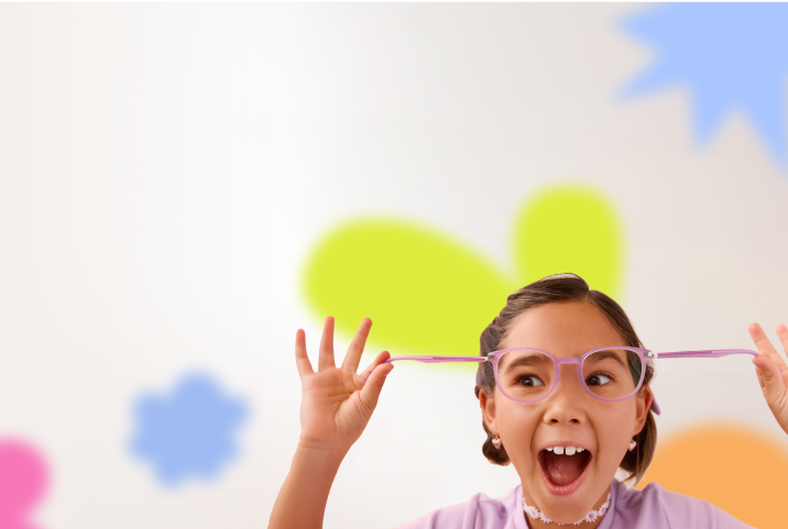 A young girl with pigtails excitedly stretches her pink glasses outward, wearing a lavender shirt and floral choker, with colorful blurred shapes in the background.