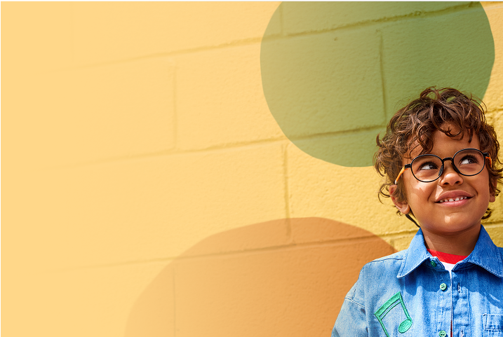 Boy with curly hair wearing round black-and-orange eyeglasses and a denim shirt, smiling while looking upward, standing against a colorful mural wall with yellow, orange, and green shapes.