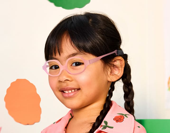 A smiling girl with long dark braids wears light pink glasses and a pink floral shirt, set against a pastel, cut-out shape background.