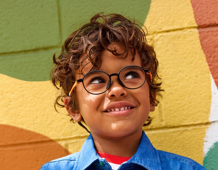 Boy with curly hair wearing round black-and-orange eyeglasses and a denim shirt, smiling while looking upward, standing against a colorful mural wall with yellow, orange, and green shapes.