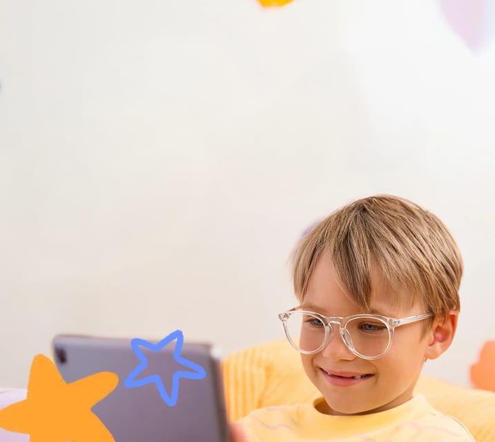 The image shows a young child with light brown hair wearing transparent glasses and a yellow sweater with pink stripes, smiling while looking at a tablet while seated on a yellow textured couch. The soft, blurred background features an orange flower shape and a blue heart shape on the wall, adding a playful touch.