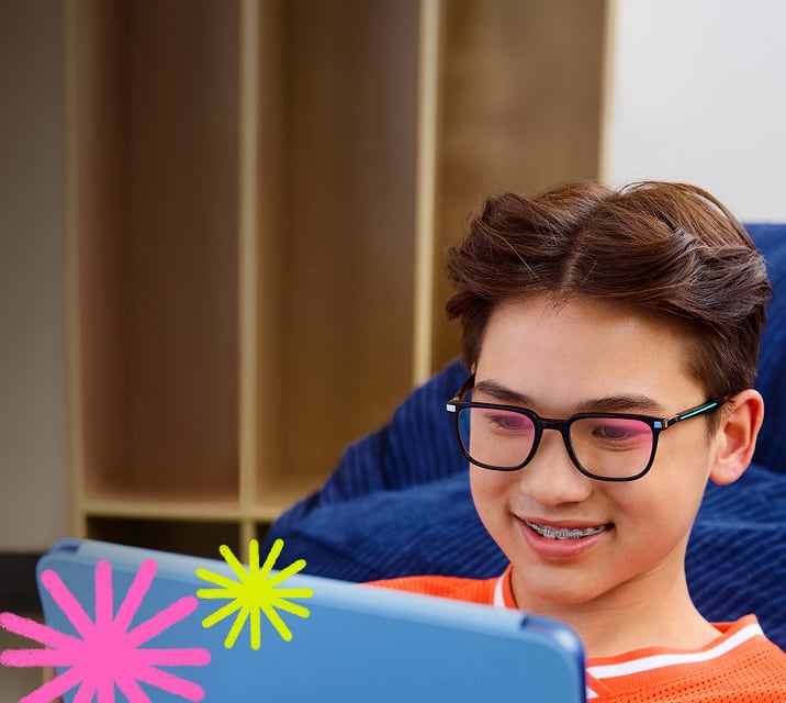 The image shows a young person with short brown hair, black rectangular glasses, and braces, smiling while looking at a blue tablet. They wear an orange shirt and sit on a blue cushion, with a blurred wall and bookshelf in the background.