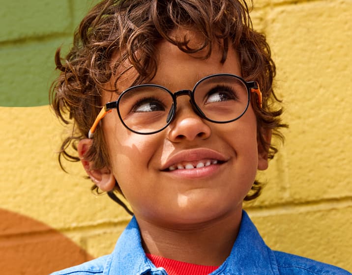 Boy with curly hair wearing round black-and-orange eyeglasses and a denim shirt, smiling while looking upward, standing against a colorful mural wall with yellow, orange, and green shapes.