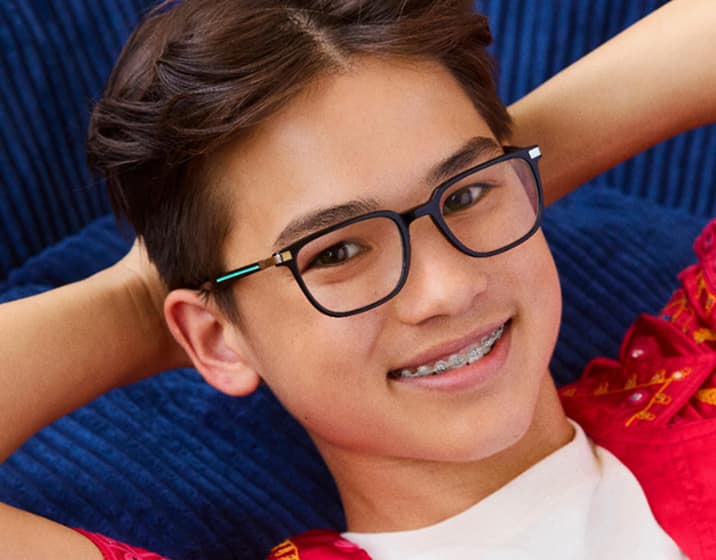 Smiling boy with short brown hair and braces wearing rectangular black-and-teal eyeglasses, a white T-shirt, and a bright pink embroidered shirt, leaning back with hands behind head against a colorful abstract background.