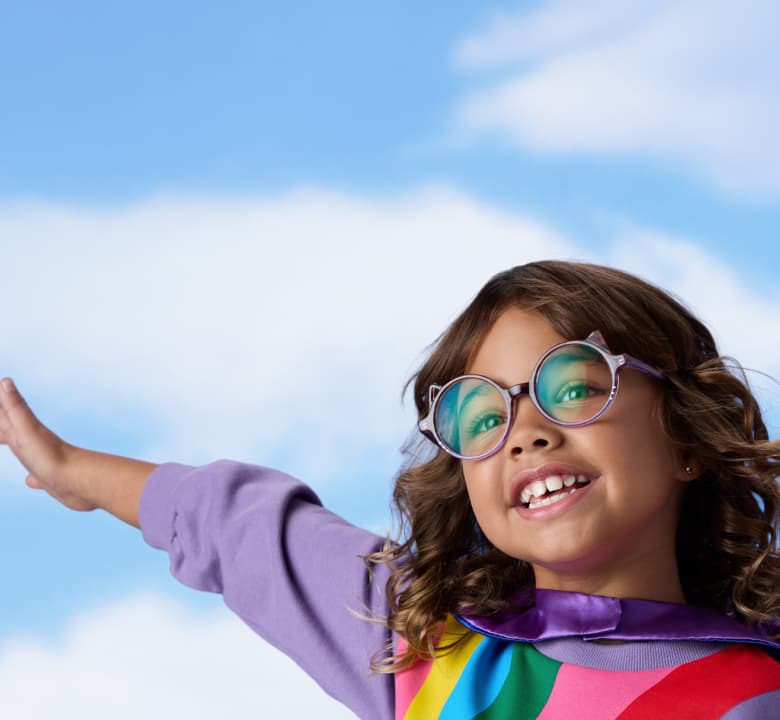 Smiling young girl wearing purple round eyeglasses and a cape extending her arm against a blue sky with soft white clouds.