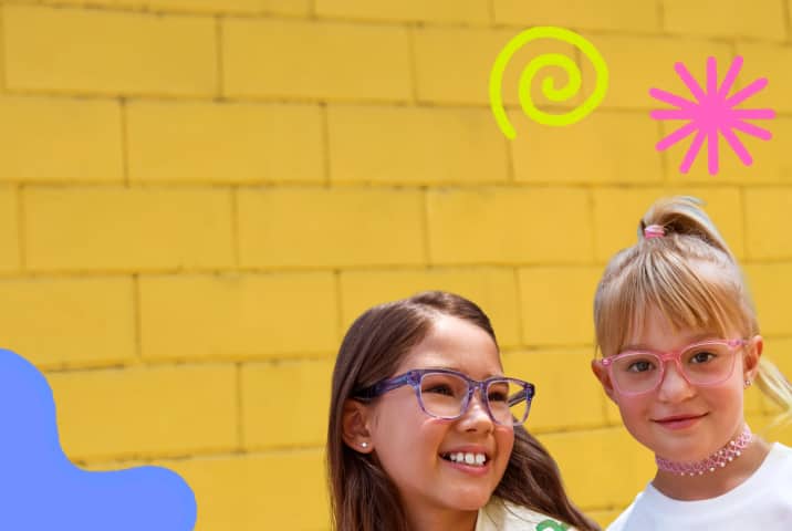 Two young girls smiling together against a bright yellow brick wall; one has blonde hair in a ponytail and wears pink rectangular glasses, while the other has dark hair and wears clear purple rectangular glasses.