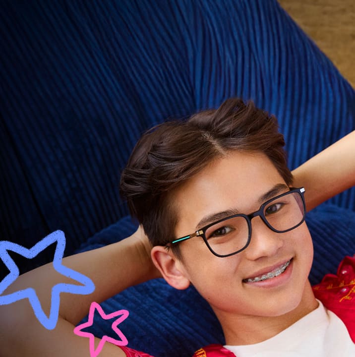 Smiling teenage boy with brown hair resting his head back in his hands, wearing rectangular black glasses with blue details on the temples, set against a dark blue corduroy background with star doodles.