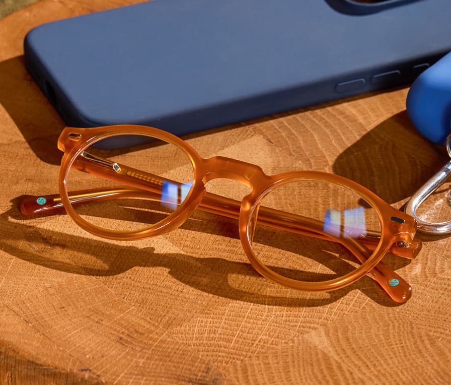 Translucent brown round Zenni glasses on a table with a phone and earbuds. Clear lenses have a slight blue reflective sheen.