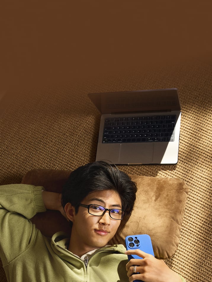 A young man laying on the ground, looking at his phone, wearing black rectangle Zenni glasses with clear Blokz lenses that have a slight blue reflective sheen.