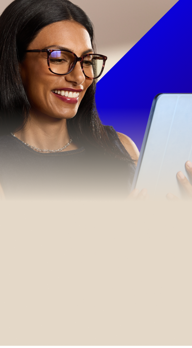 Smiling woman with long dark hair wearing tortoiseshell glasses while looking down at a tablet against a warm beige background.