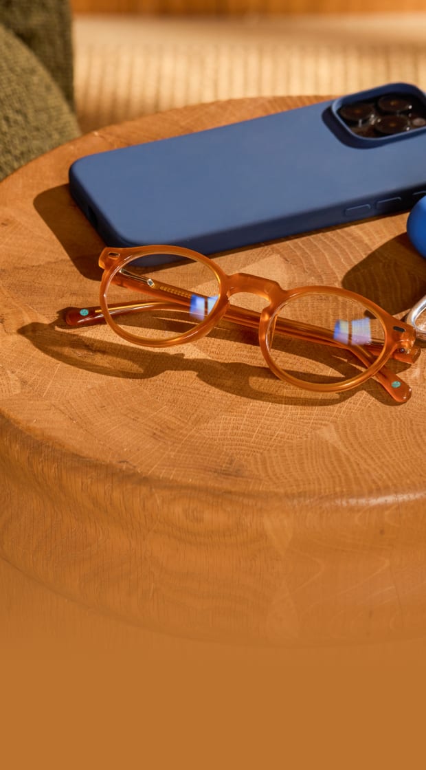 Translucent orange round eyeglasses resting on a wooden side table next to a blue smartphone.