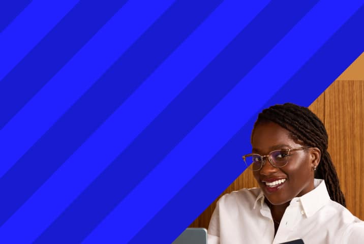 Smiling woman wearing clear wireframe eyeglasses and a white button-down shirt looking toward the camera, set against diagonal royal blue stripes and a warm wood background.