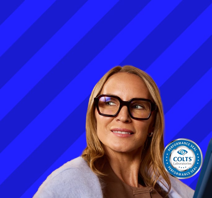 Close-up of a smiling blonde woman looking to the side while wearing thick, square, dark brown eyeglasses, set against a dark blue background with diagonal stripes, featuring a 'COLTS Laboratories Performance Seal' icon in the lower right.