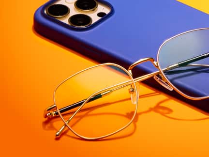 A pair of gold-framed, blue light-filtering glasses rests on top of a blue smartphone case against a vibrant orange background. The lenses reflect a subtle blue tint, and the temples feature a mix of gold and black detailing.