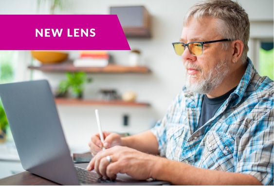 Image of a man sitting at a desk wearing Zenni glasses with blokz lenses in them, working on a computer with a note that says ‘new lens’. 