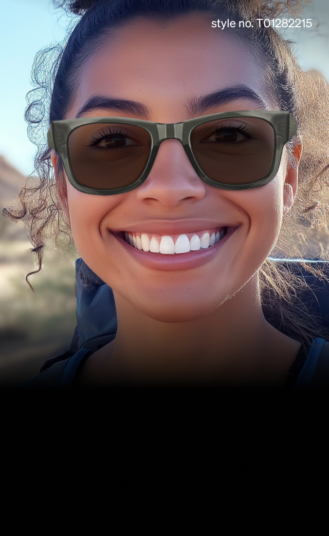 A woman smiles in the desert while wearing green, military-style sunglasses. The sun shines brightly in the background, with distant mountains and a tree adding to the natural, rugged setting.