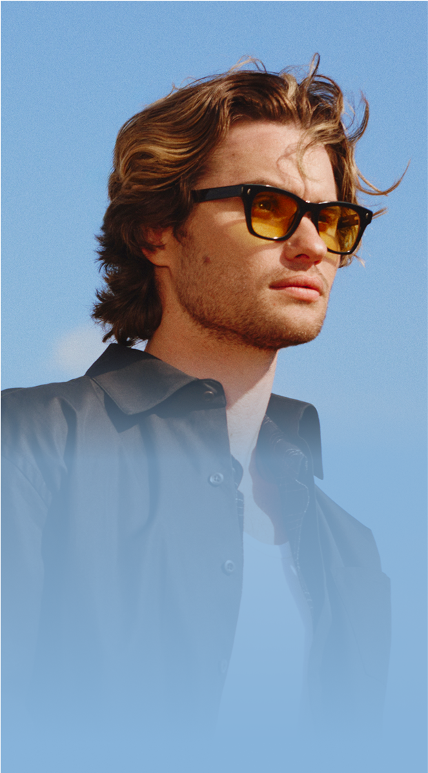 Chase Stokes wearing Zenni black frames with tinted lenses, standing outside with a clear blue sky in the background.