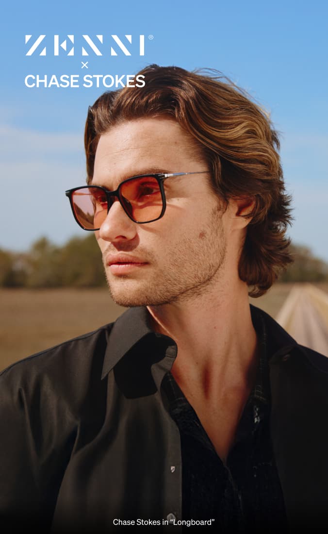 Chase Stokes wearing gradient tinted sunglasses 'Longboard' and a black collared shirt looks off to the side against a scenic backdrop of an open road, clear blue sky, and distant greenery.