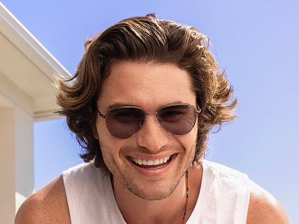Chase Stokes wearing sunglasses leans forward in a white tank top under a clear blue sky.